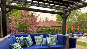 A covered outdoor seating area with a blue sofa and cushions, a fire pit, and red umbrellas. - 9 Bedroom Historic Summit Mansion With Pool And Hot Tub (St Paul)