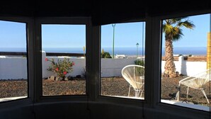 Una habitación con vistas al mar a través de grandes ventanales, una silla blanca y un pequeño jardín con una palmera y flores.