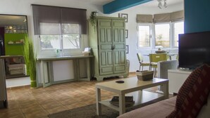 Una sala de estar con sofá, mesa de centro, televisor, una zona de cocina con armarios y una ventana, además de una planta verde.