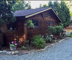 Cozy cabin with a sloped roof and a small porch area, surrounded by lush gardens and a gravel driveway.