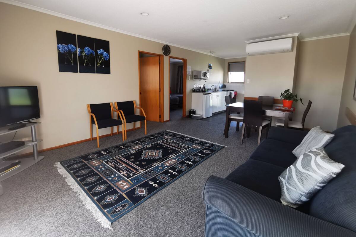Una sala de estar con sofá, televisión, comedor con sillas, cocina y un pasillo.