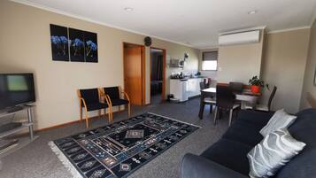 A living room with a sofa, a TV, a dining area with chairs, a kitchen, and a hallway.