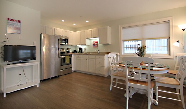A modern kitchen with white cabinets, a stainless steel refrigerator, a microwave, and an oven. There is a dining table with white chairs and a window with blinds.