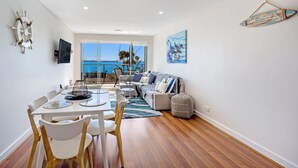 Un salon moderne avec un balcon donnant sur un plan d’eau, un coin repas avec une table et des chaises, et un espace détente avec un canapé et des coussins.