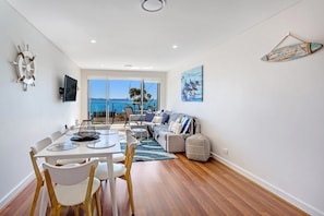 Een moderne woonkamer met een balkon met uitzicht op een waterpartij, een eethoek met een tafel en stoelen, en een zithoek met een bank en kussens.