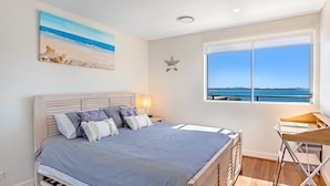 Une chambre à coucher avec un lit, un bureau, une fenêtre donnant sur la mer et un tableau accroché au mur.