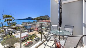 Un balcon doté d’une table et de chaises en verre, offrant une vue sur une zone côtière avec des bâtiments et des bateaux.