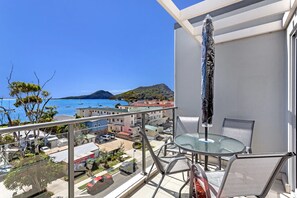 Een balkon met een glazen tafel en stoelen, met uitzicht op een kustgebied met gebouwen en boten.