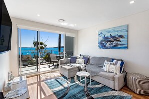 A modern living room with a large window, a sofa set, a coffee table, and a painting of dolphins.