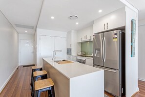 A modern kitchen with a stainless steel refrigerator, a built-in oven, and a white countertop. There are three wooden stools and a sink with a modern faucet.