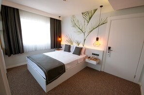 Una habitación de hotel moderna con una cama grande, una mesita de noche con lámpara, una planta en la pared y una puerta con cerradura electrónica.