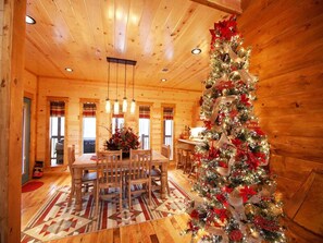 Interior acogedor de una cabaña de troncos, con comedor que incluye una mesa y sillas de madera, un árbol de Navidad y una alfombra estampada.