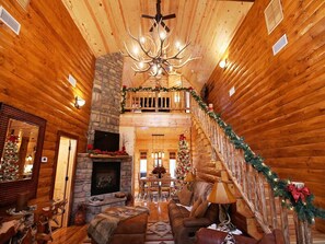 Interior de una cabaña de troncos con chimenea de piedra, escalera de madera y adornos navideños.