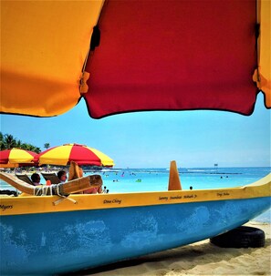 Un bote con casco amarillo y azul, llamado "Dino Ching", atracado en una playa con gente y sombrillas al fondo.