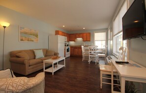 A living room with a sofa, coffee table, and a TV. There is a kitchen with wooden cabinets and a dining area with a table and chairs.