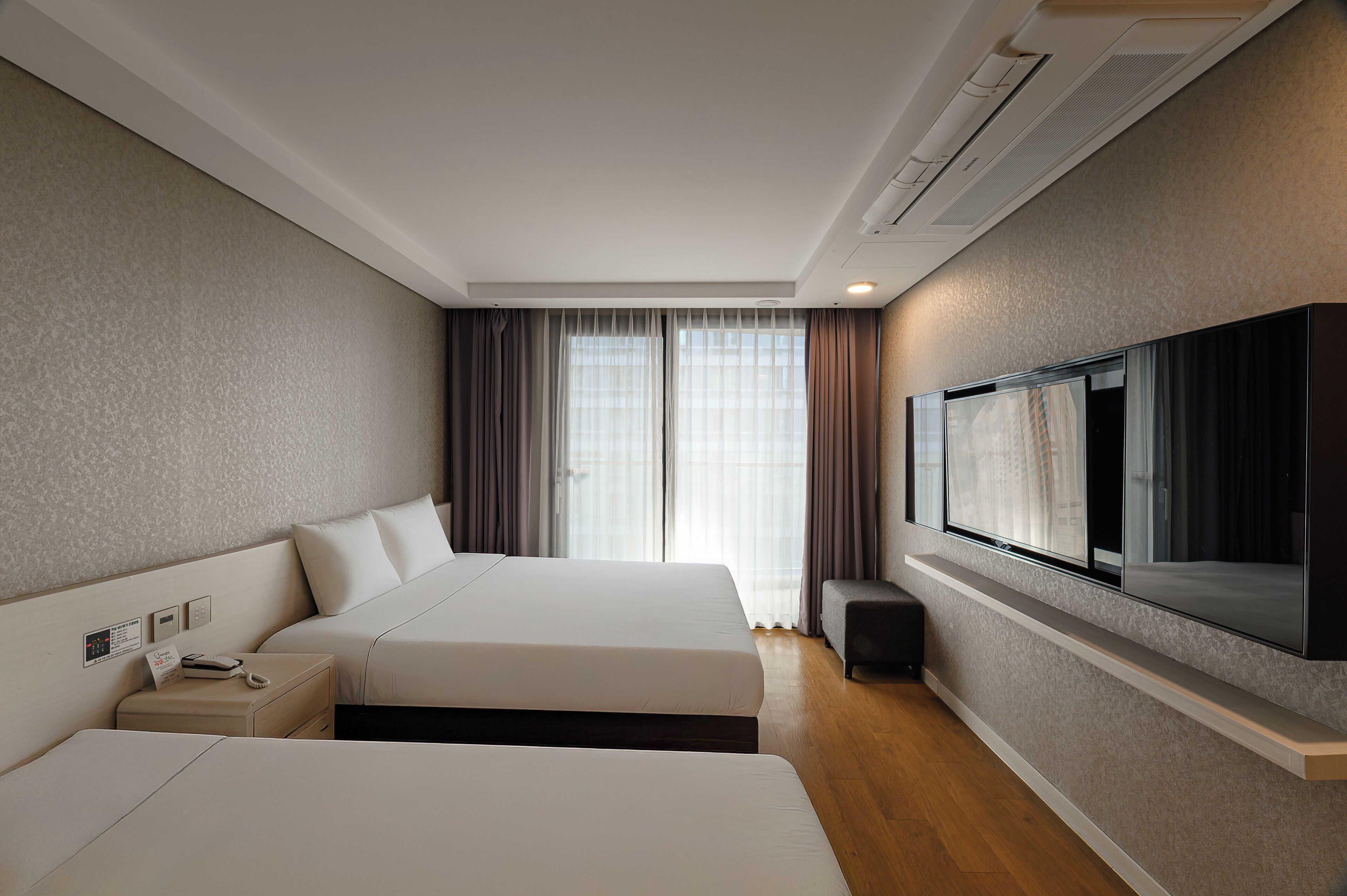 A hotel room with two beds, a TV mounted on the wall, a bedside table with a phone, and a window with curtains.