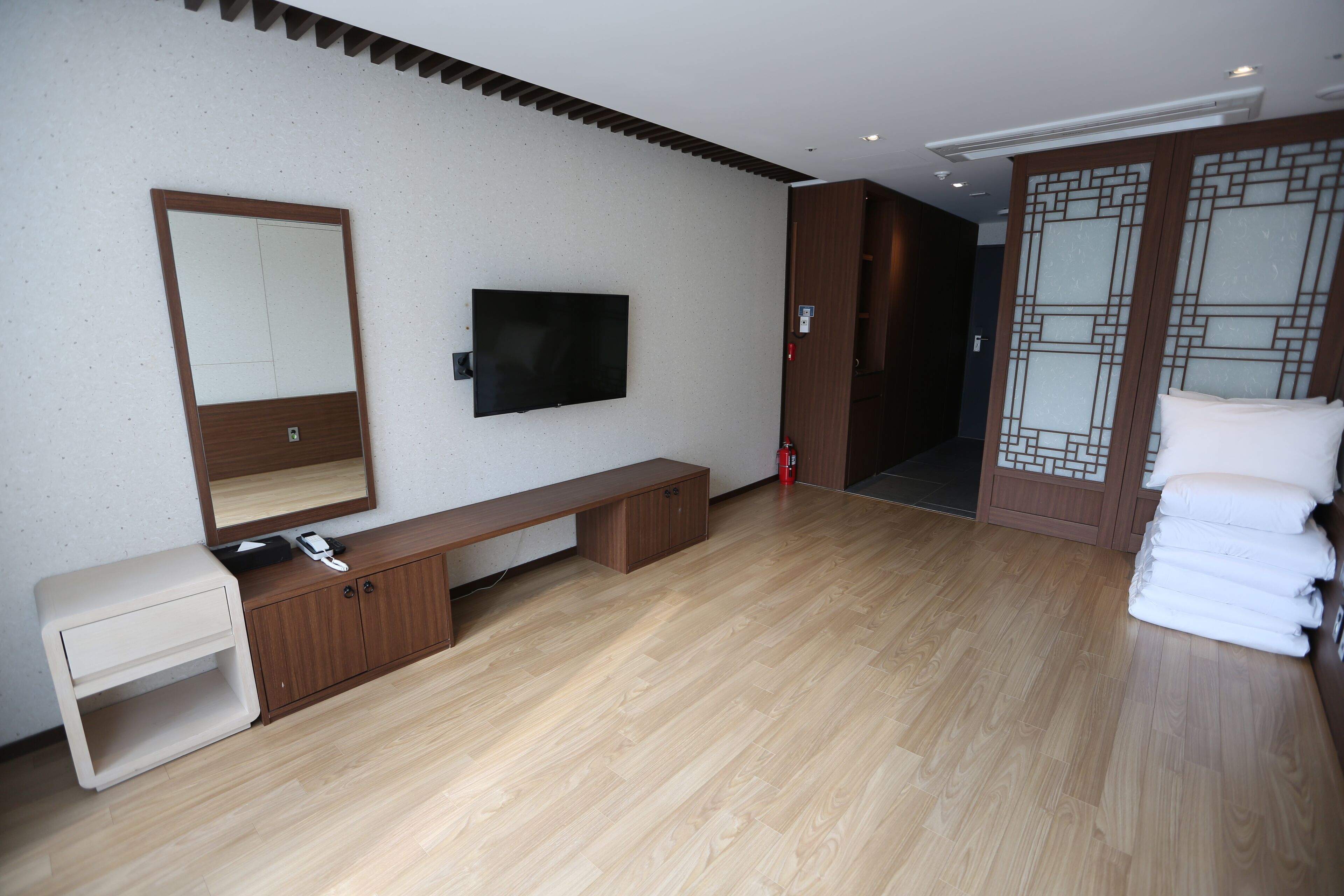 A modern hotel room with a wooden floor, a large mirror, a flat-screen TV, a bench, a small table, and a stack of towels.