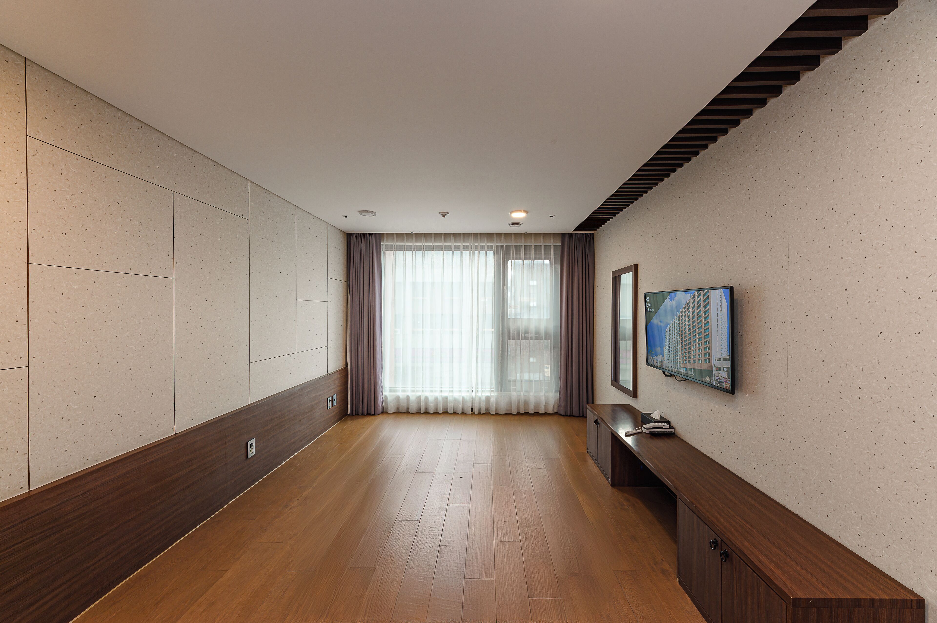 Un pasillo de hotel con suelo de madera, un banco de madera, un televisor de pantalla plana montado en la pared y un ventanal con cortinas translúcidas.