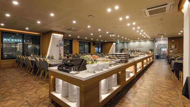 Un buffet avec plusieurs points de service, un long comptoir où sont présentés les plats, et un espace repas avec des tables et des chaises.