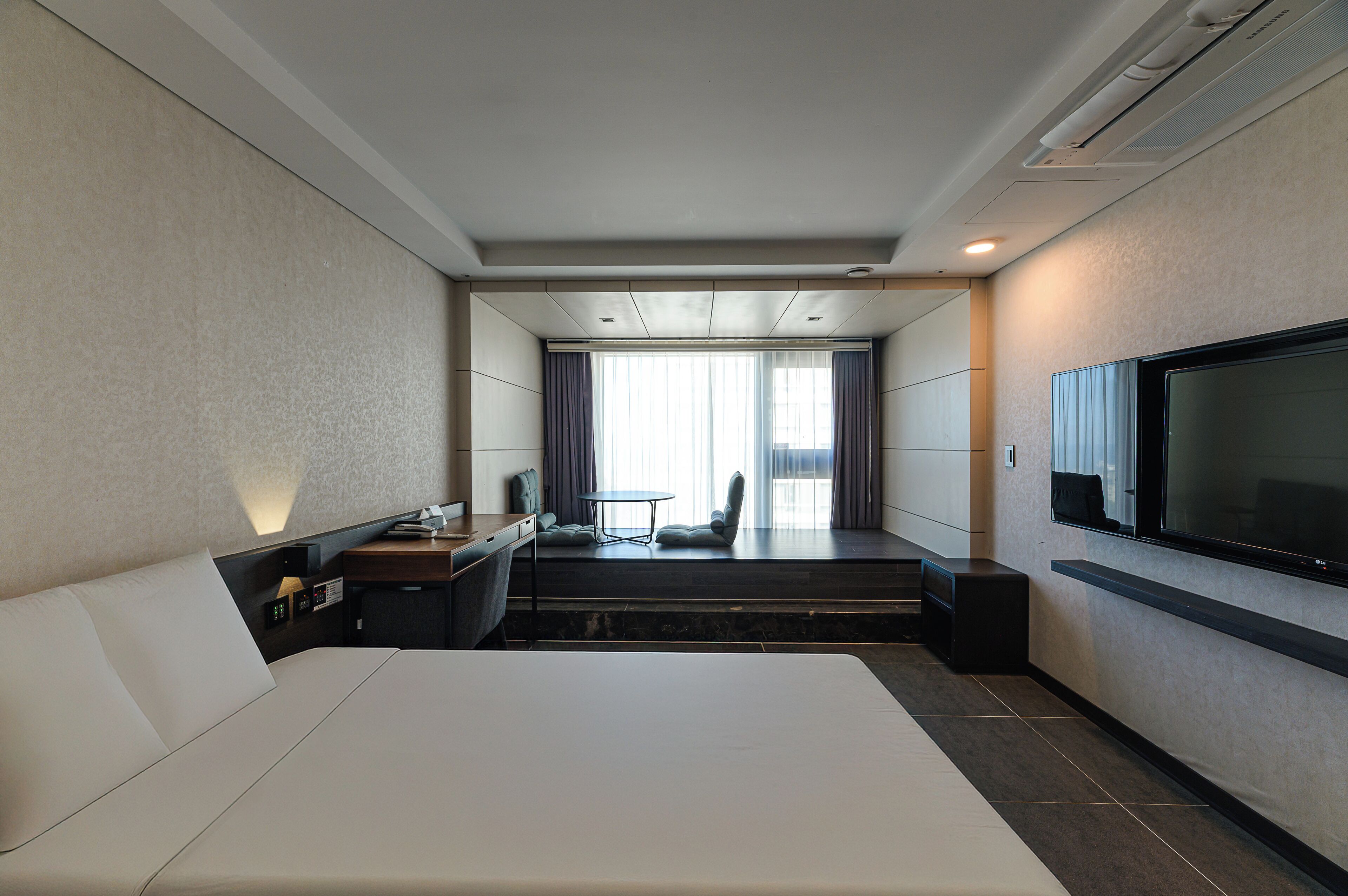 Ein Hotelzimmer mit einem großen Bett, einem Schreibtisch mit Stuhl, einem an der Wand montierten Fernseher und einem Fenster mit Vorhängen.
