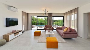 Un salon moderne doté d’un téléviseur à écran plat, de deux canapés, d’une table basse et d’une grande fenêtre offrant une vue sur la verdure et une piscine.