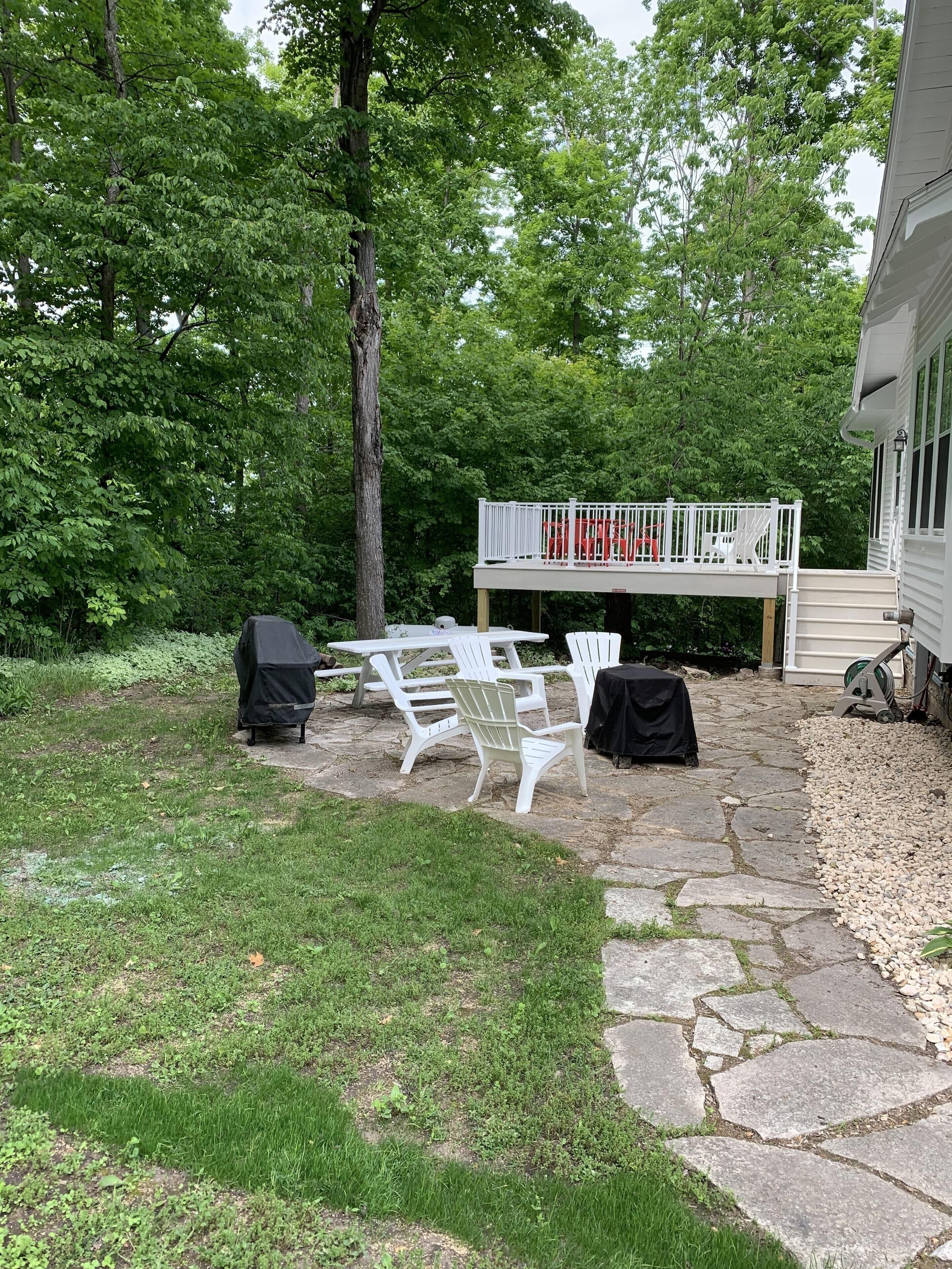 A backyard with a stone patio, white plastic chairs, a covered grill, and a deck with a railing.