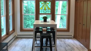 Comedor con una mesa y cuatro sillas, ventanas amplias, ventilador de techo y vista a zonas verdes exteriores.