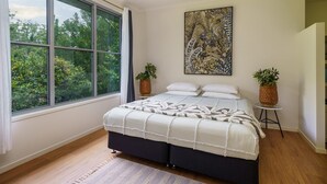 Ein Schlafzimmer mit großem Fenster, einem Bett mit gemustertem Überwurf, zwei Blumentöpfen und einem an der Wand hängenden Kunstwerk.