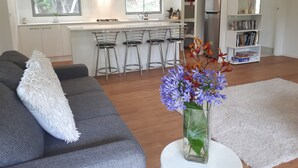 Ein modernes Wohnzimmer mit einem grauen Sofa, einem weißen Zierkissen, einer Vase mit Blumen und einem weißen Teppich. Im Hintergrund befindet sich eine Küche mit weißen Schränken, eine Bar mit Metallstühlen und ein Bücherregal mit Büchern und Dekorationsobjekten.