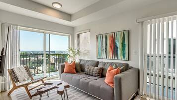 Un salon moderne avec un canapé gris, des coussins orange, une chaise en bois et une grande fenêtre donnant sur la ville.