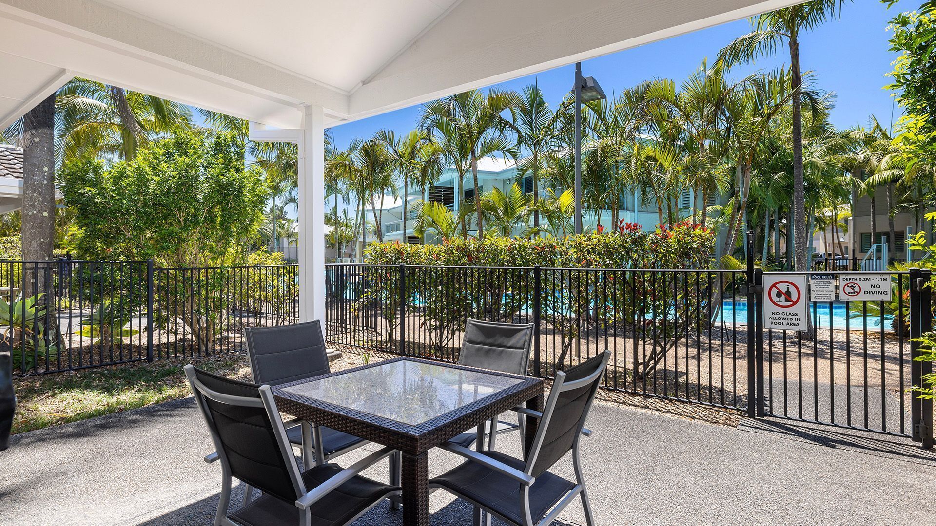 An outdoor seating area with a glass table and chairs, surrounded by a metal fence, overlooking a pool and palm trees.