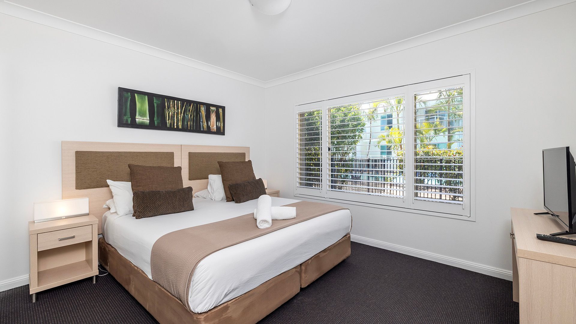 A hotel room with a bed, bedside tables, a TV, and a window with blinds.