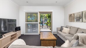 Ein modernes Wohnzimmer mit Flachbildfernseher, Couchtisch aus Holz, grauem Sofa und Blick durch eine Schiebetür auf eine begrünte Außenanlage.