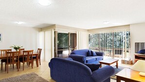 Una sala de estar con un sofá azul, una mesa de comedor de madera y sillas, un televisor y un ventanal corredero que da a un balcón.