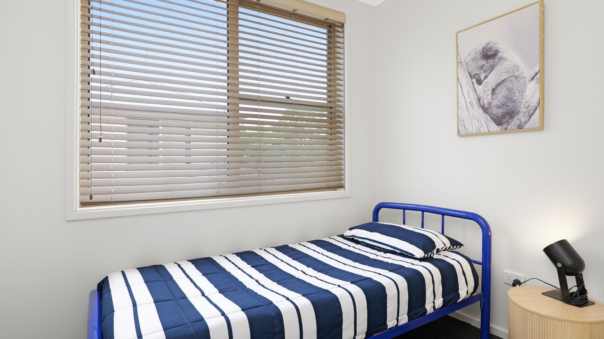 A single bed with a blue metal frame, a striped bedspread, and a bedside lamp on a wooden nightstand.