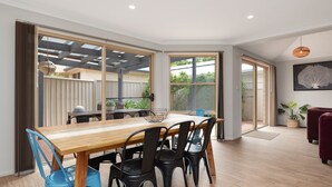 Een eetgedeelte met een houten tafel en stoelen, grote schuifdeuren naar een achtertuin, en een rode bank in de woonkamer.