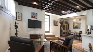 Un salon équipé d’un téléviseur, de deux fauteuils en bois, d’une table basse et d’un tapis à motifs. Un escalier mène à une salle à manger où se trouvent une table et des chaises.