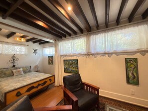 Un dormitorio con cama, una silla, un cuadro y una ventana con cortinas.