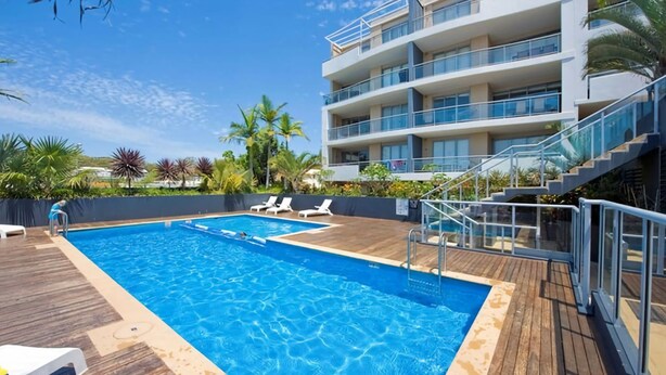 A swimming pool with a wooden deck, lounge chairs, and a multi-story building with balconies. - Cote D Azur in the heart on Nelson Bay with a swimming pool (Nelson Bay)
