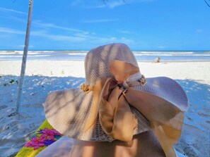 A beach scene with a person lying on a blanket, a beach chair, and a clear view of the ocean. - FERIASMARES – Beachfront House in Ilhéus, until 10. (Ilhéus)