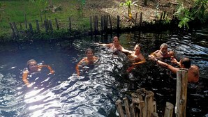 Um grupo de pessoas em uma piscina natural, cercada por estacas de madeira e vegetação.