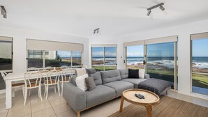 Ein modernes Wohnzimmer mit einem grauen Modulsystem, einem runden Holztisch und einem Essbereich mit weißen Stühlen. Große Glastüren führen zu einem Balkon mit Meerblick.