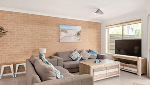 A living room with a brick wall, a sofa, a coffee table, a TV, and a window with a view. - Walking Distance to Little Beach and Shoal Bay (Nelson Bay)