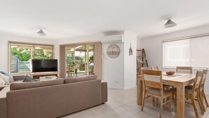 A modern living room with a sofa, TV, and dining area with wooden chairs and a table. - Walking Distance to Little Beach and Shoal Bay (Nelson Bay)