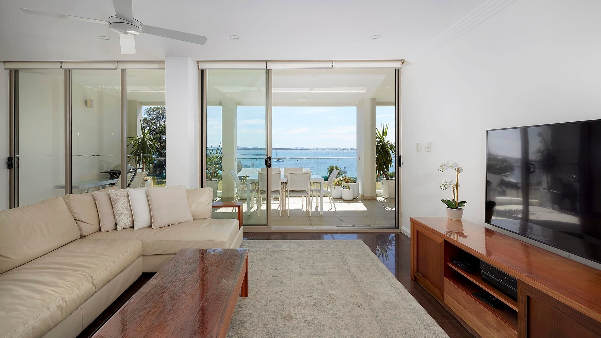 Een moderne woonkamer met een beige bank, een houten salontafel, een flatscreen televisie en een schuifdeur naar een balkon met uitzicht op zee.