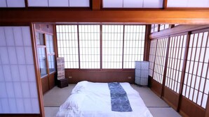 Una habitación tradicional japonesa con suelo de tatami, una cama con colcha blanca y azul, y puertas correderas shoji en las ventanas.
