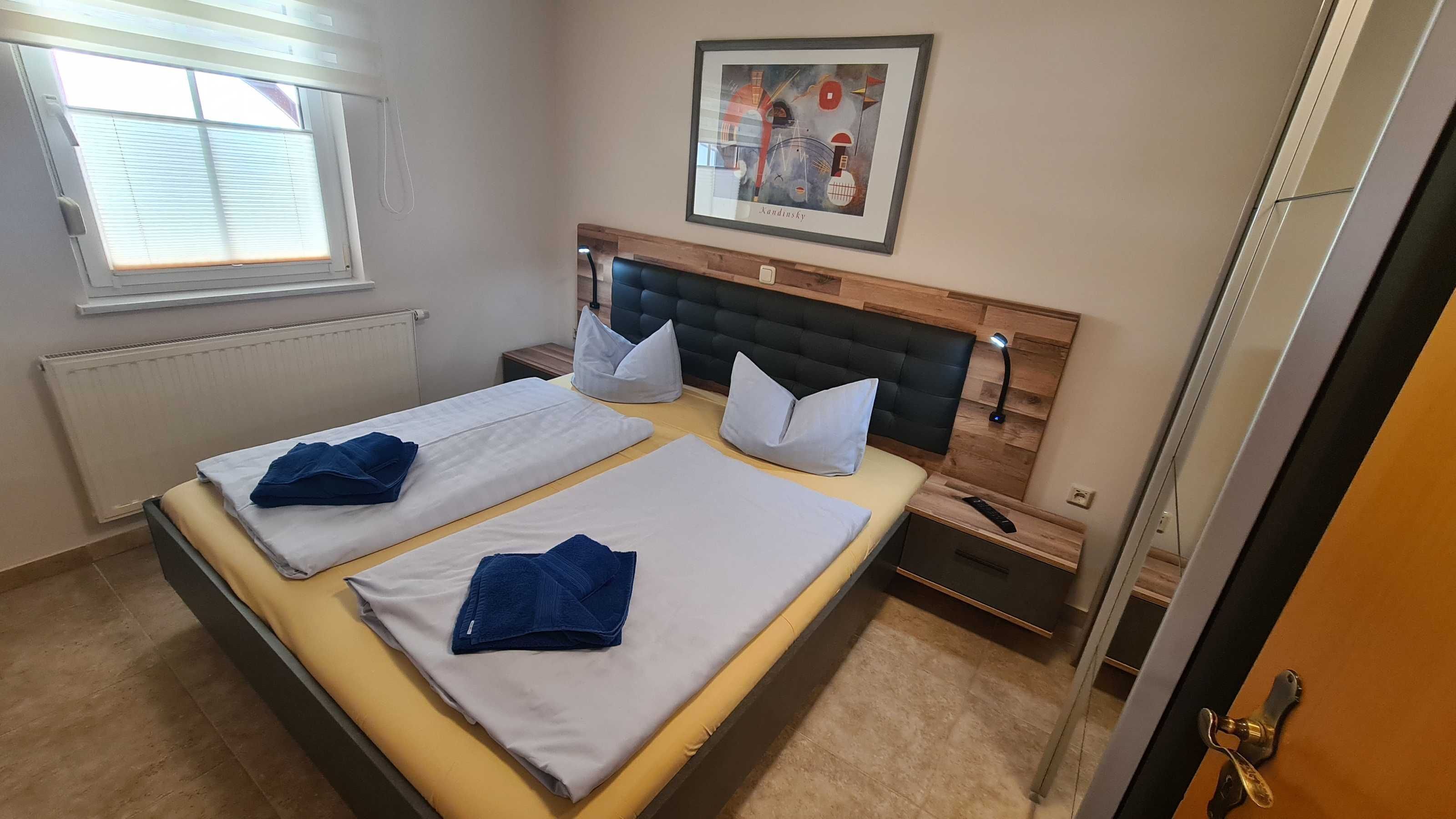 Uma cama de casal com duas almofadas e um cobertor, uma mesa de cabeceira com abajur, uma janela com persianas e um quadro na parede.