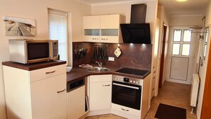 Una cocina compacta con microondas, estufa, horno y un lavamanos pequeño. Hay armarios, una ventana con persianas y una puerta que conduce a otra habitación.