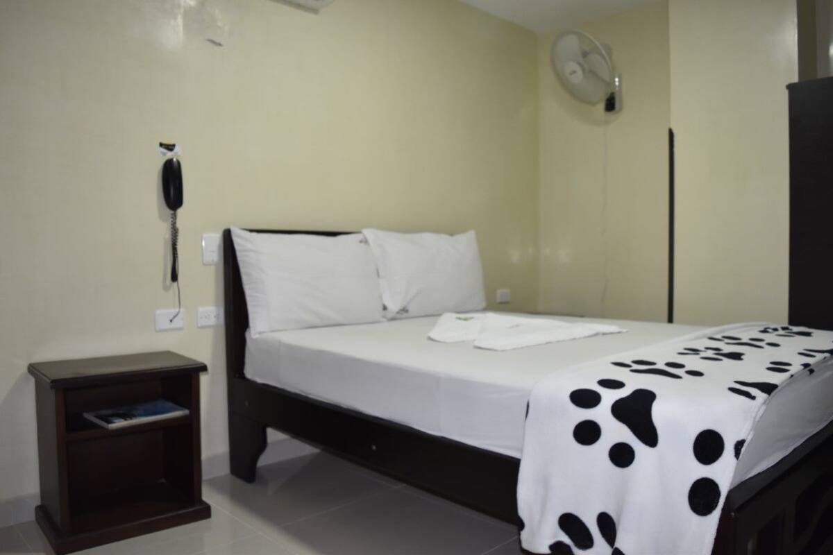 Una habitación de hotel con cama, mesita de noche, teléfono en la pared y ventilador de pared.