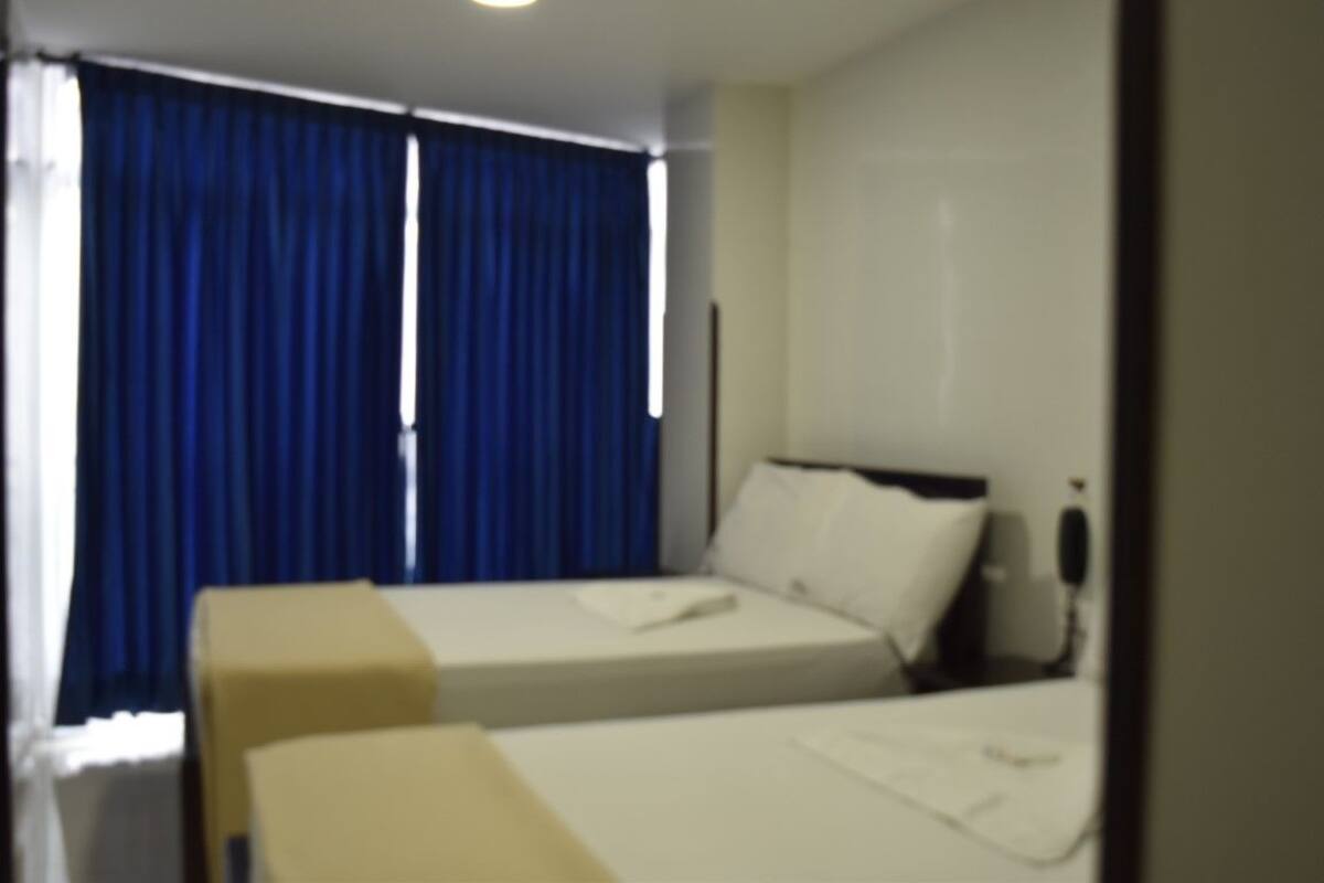 Habitación de hotel con dos camas, una ventana con cortinas azules y una mesita de noche con lámpara.